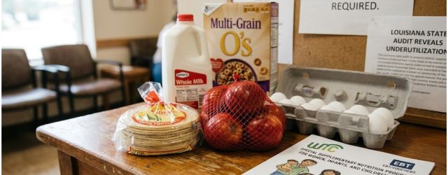 Des aliments essentiels WIC inutilisés, posés sur une table d'information dans une clinique, avec un panneau d'affichage signalant les contraintes d'accès